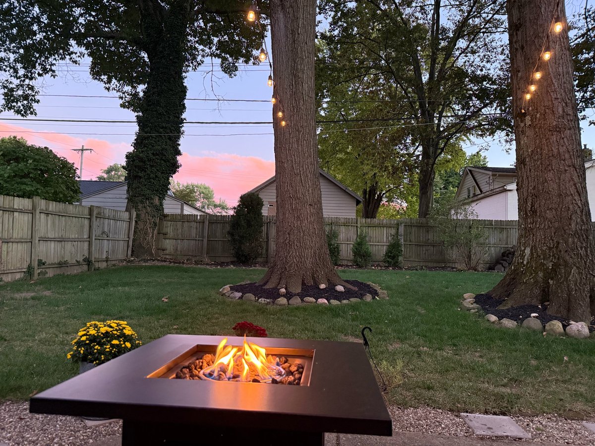 A modern fire pit glowing in a backyard at dusk with tall trees, yellow flowers, and residential homes visible in the background