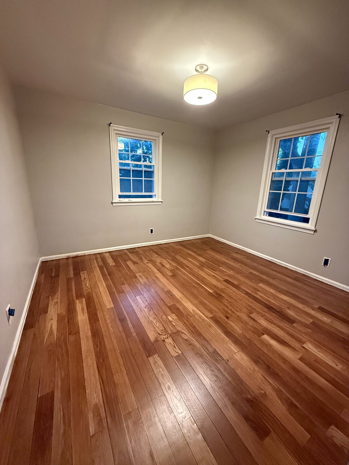 Empty room with hardwood floors, white walls, two windows with blue night sky, and ceiling light fixture