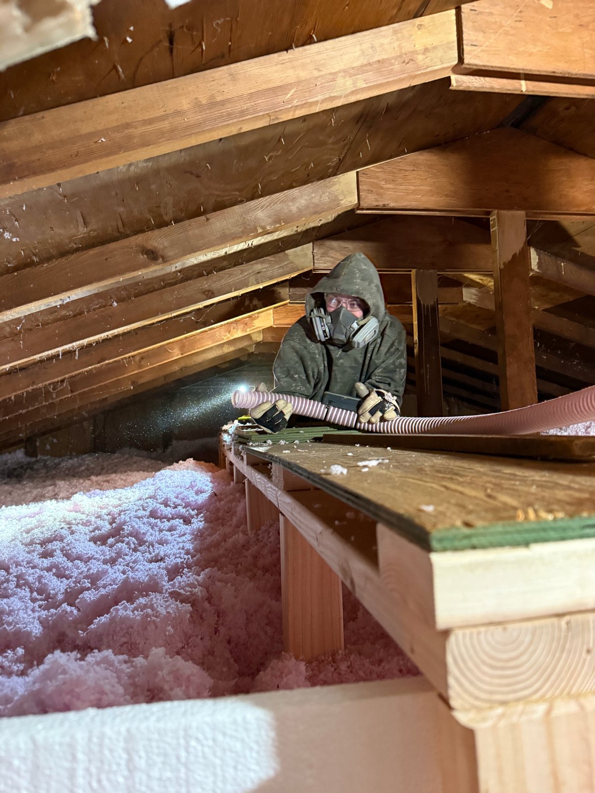 Worker in protective gear installing insulation in attic space between wooden beams