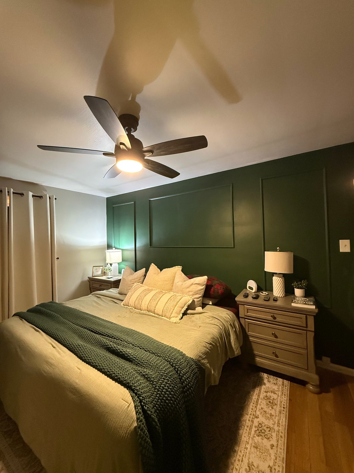Modern bedroom with dark green accent wall, queen bed with yellow pillows, ceiling fan with light, and wooden nightstand with lamp